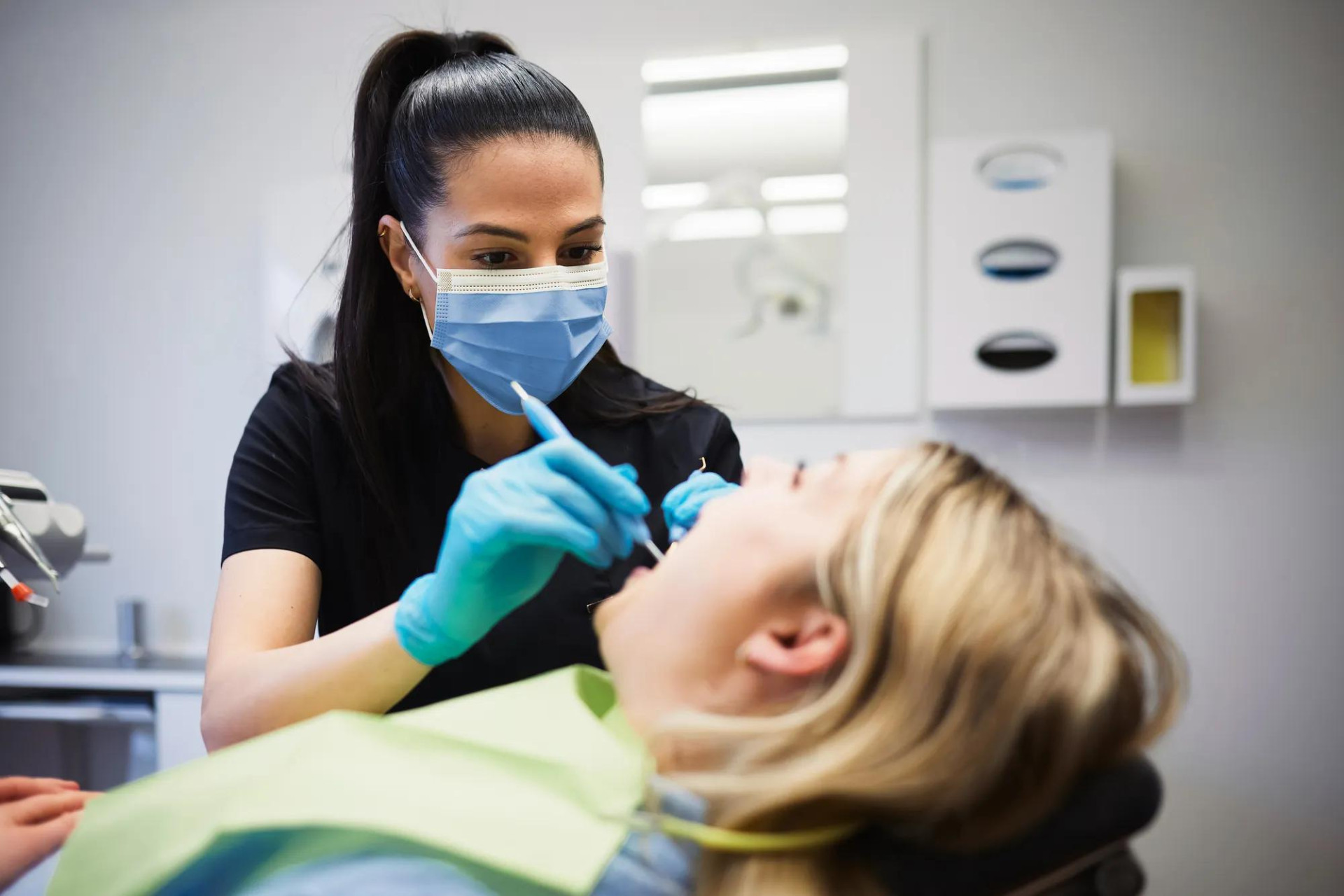 A dental professional wearing a mask and gloves cleans a patient’s teeth while the patient reclines in a dental chair.