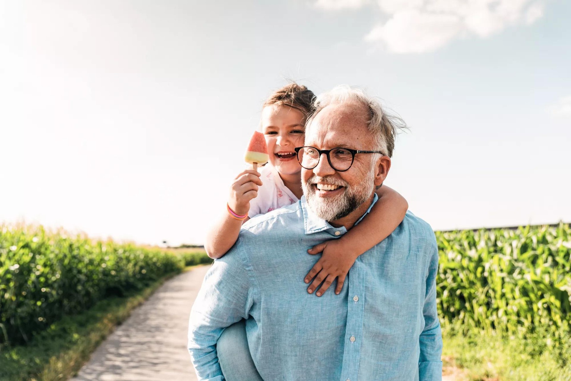 Father joyfully carries his young child on his back through a field, while the child enjoys a popsicle. Father joyfully carries his young child on his back through a field, while the child enjoys a popsicle.