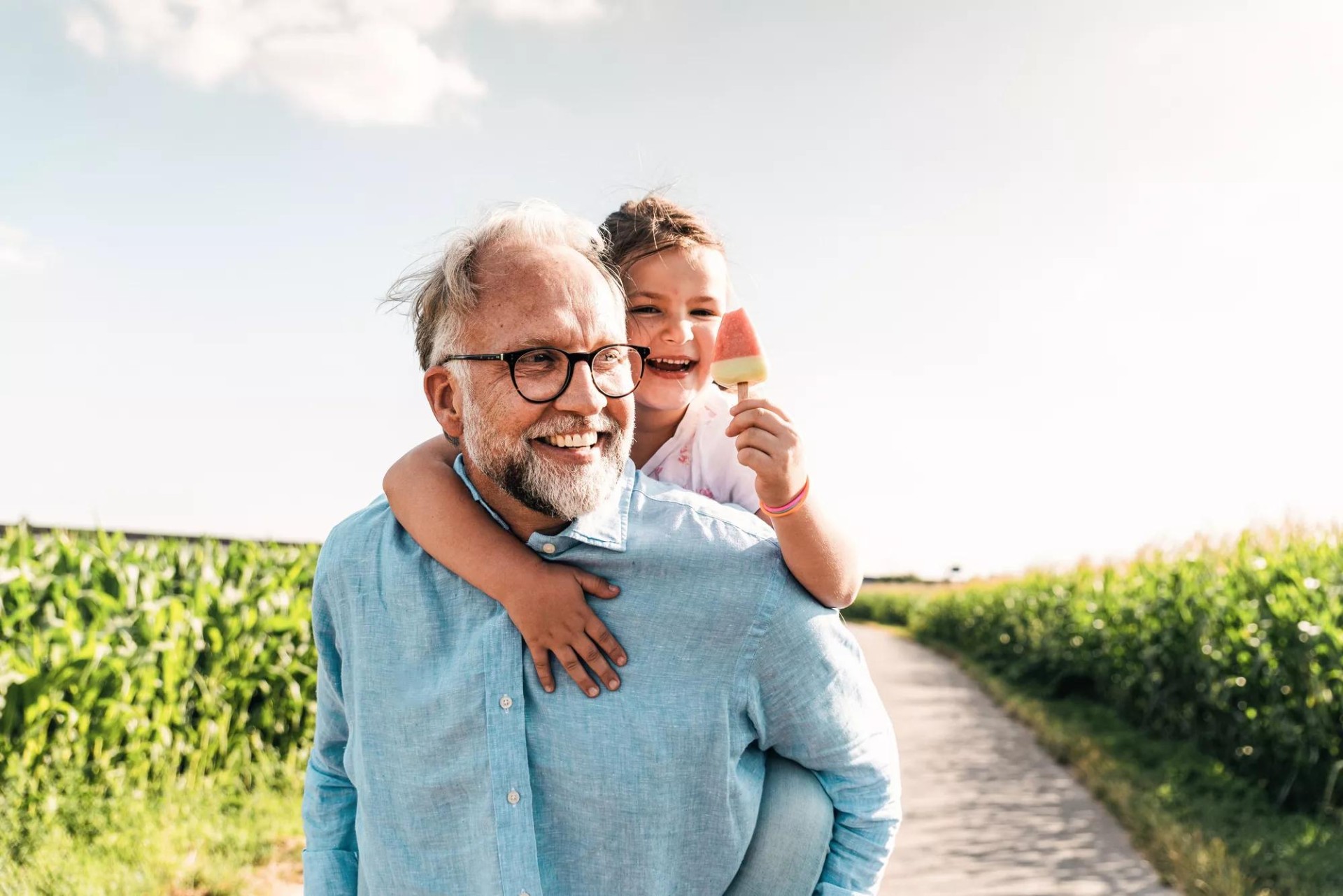 Father joyfully carries his young child on his back through a field, while the child enjoys a popsicle. Father joyfully carries his young child on his back through a field, while the child enjoys a popsicle.